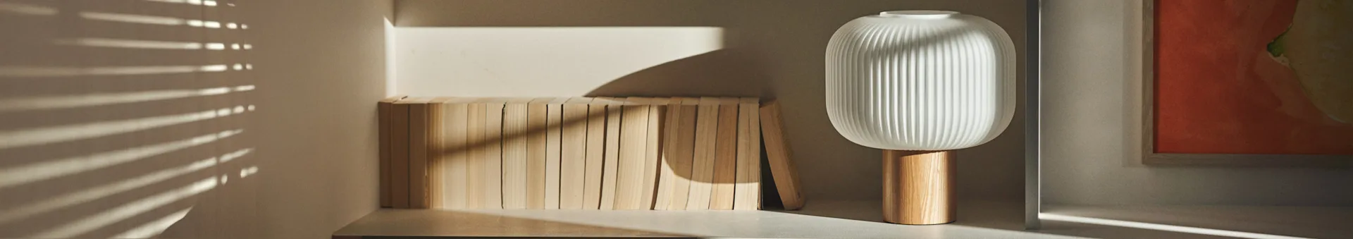 Une lampe moderne blanche nervurée avec une base en bois se dresse à côté d'une rangée de blocs de bois clair, la lumière du soleil projetant des ombres horizontales.