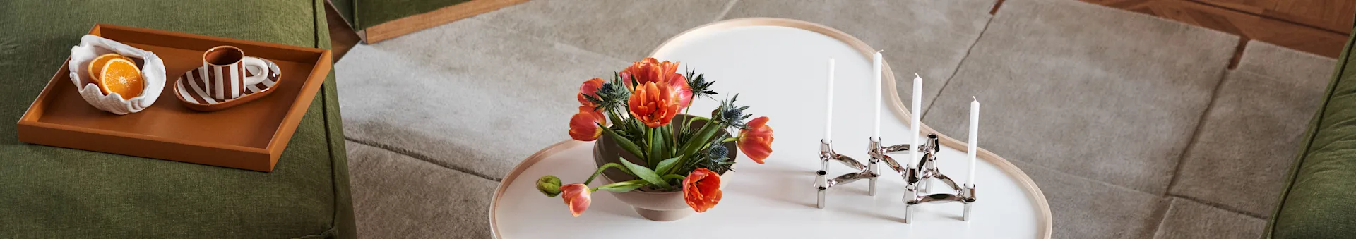Canapé vert, table basse avec tulipes oranges et chandelier en argent. Plateau sur le canapé avec des tranches d'orange et une tasse rayée.