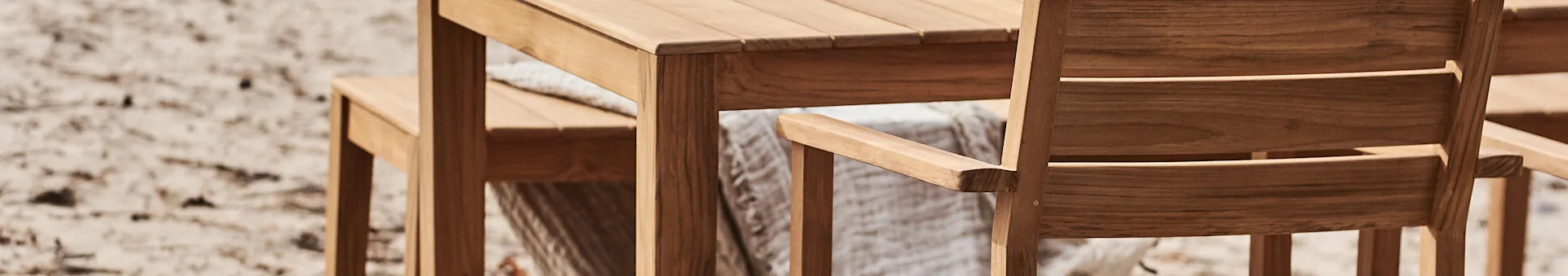 Gros plan d'une table et de chaises de jardin en bois, avec un plaid clair drapé sur une chaise, sur un fond sableux flou.