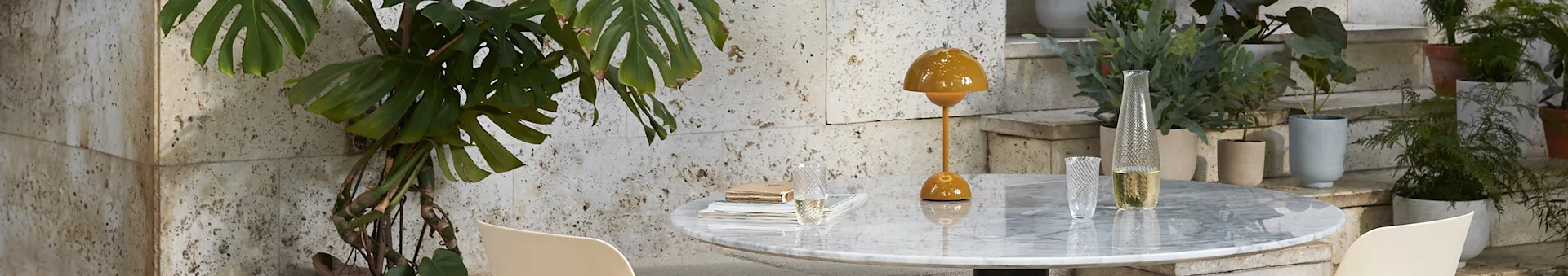 Lampe jaune moutarde et vase en verre sur table en marbre, avec monstera à gauche et plantes en pot sur escaliers à droite.