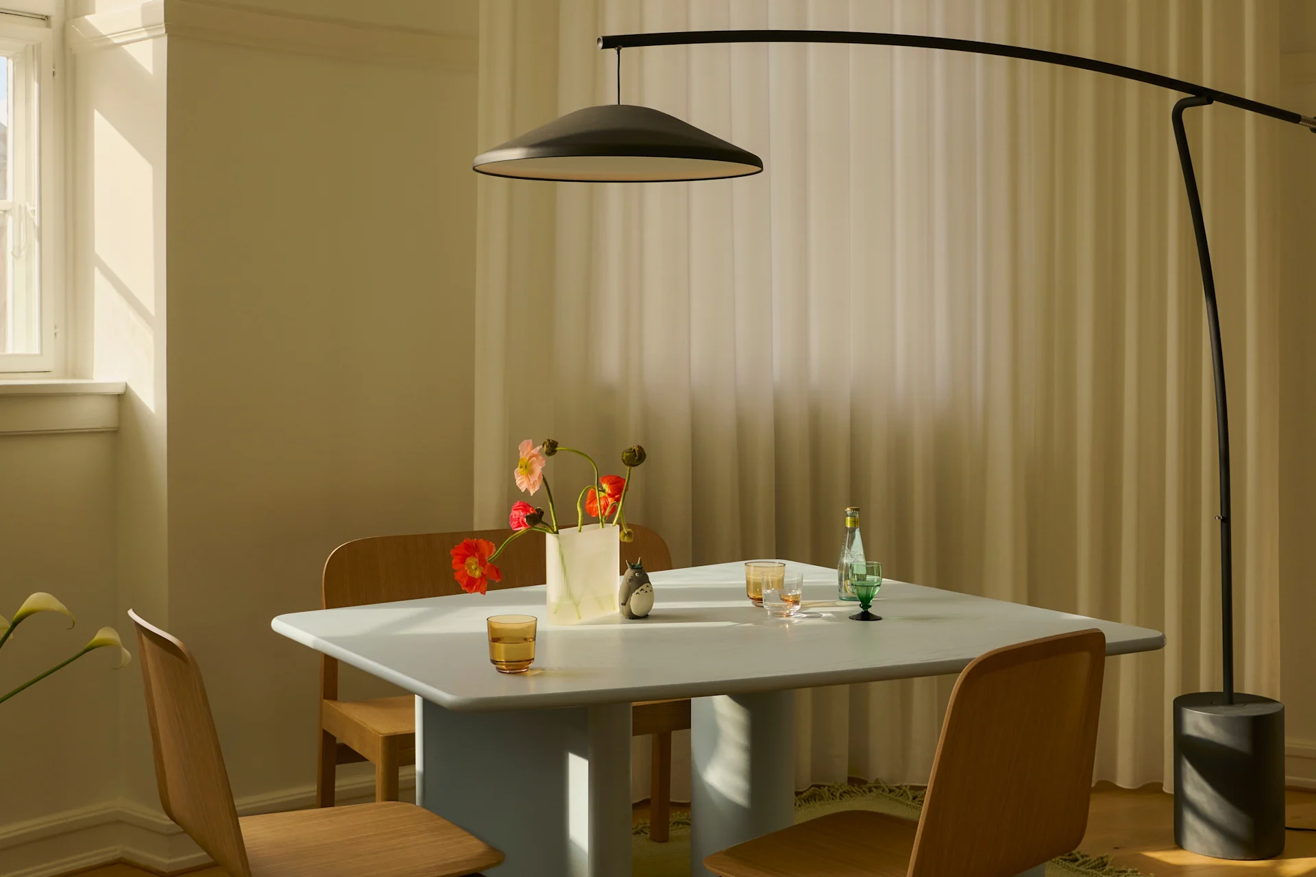 Salle à manger moderne avec table bleue claire, chaises en bois, lampadaire noir et vase de fleurs colorées au soleil.