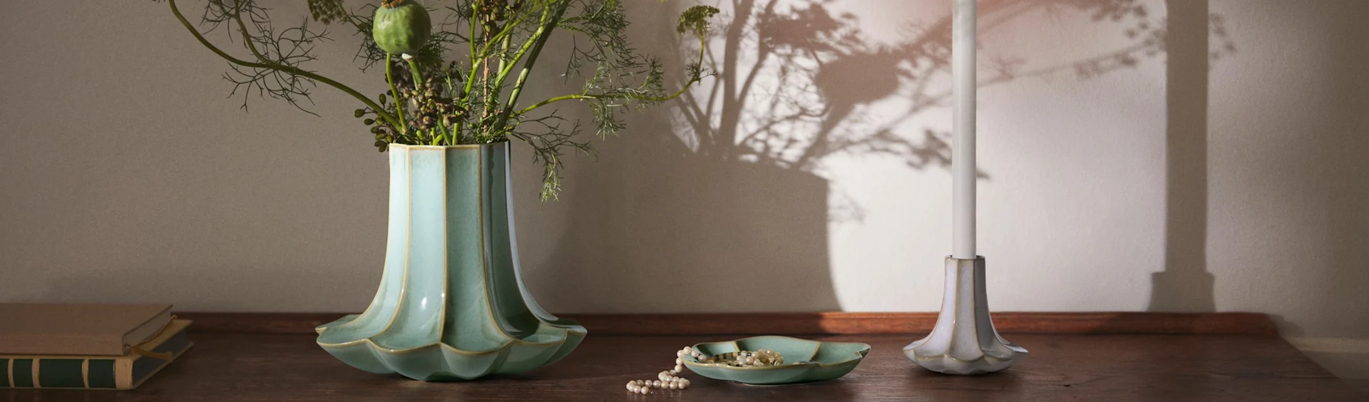 Un vase cannelé bleu sarcelle avec du feuillage, un collier de perles dans un plat assorti, une bougie blanche dans un bougeoir cannelé et des livres sur une surface en bois.