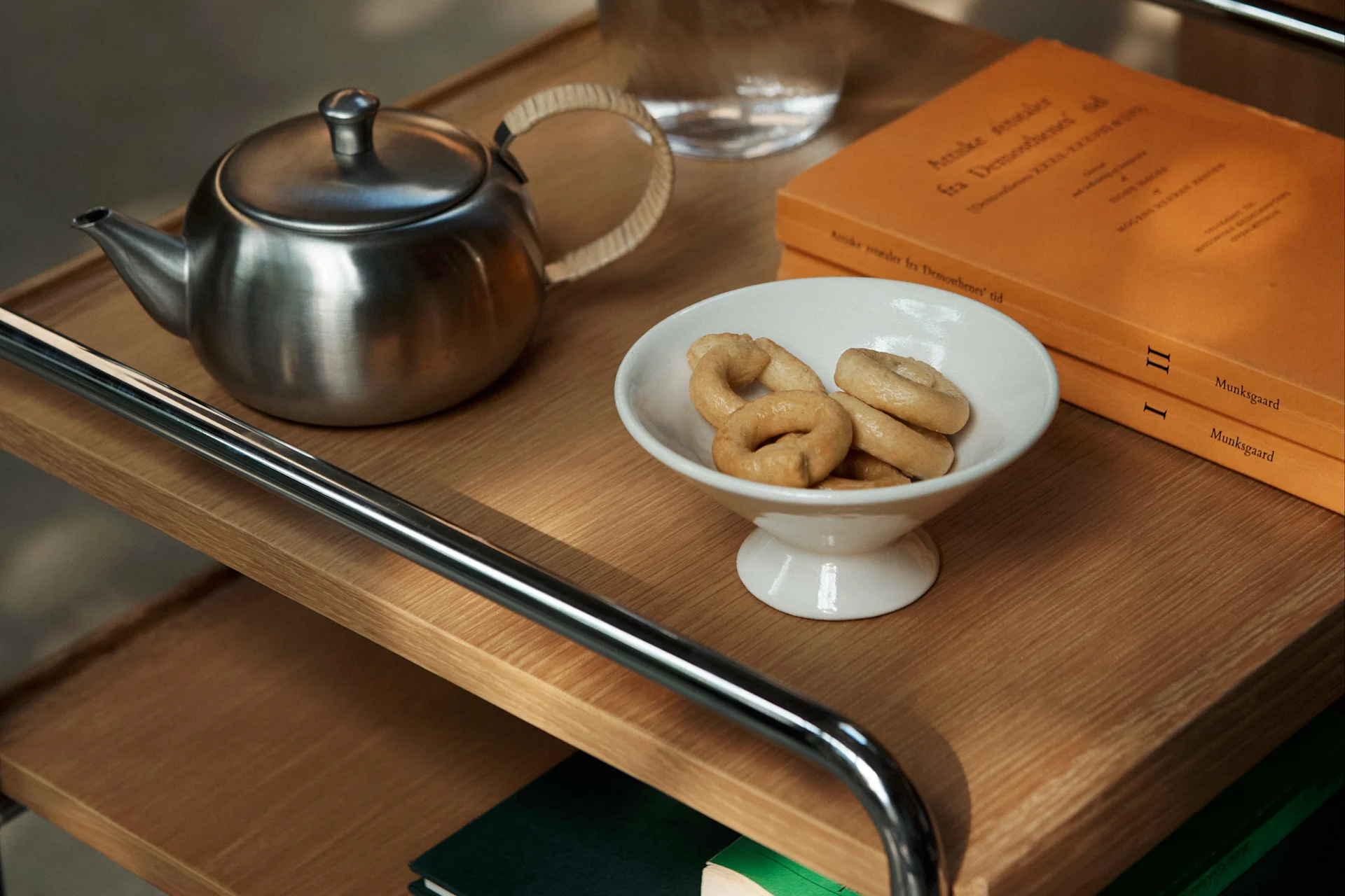 Une théière en métal, un bol blanc de biscuits en anneau et deux livres oranges sur un chariot en bois.