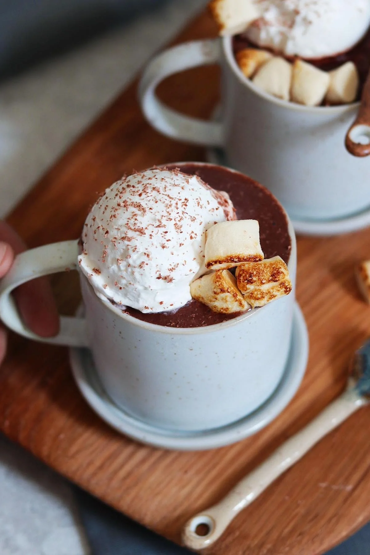 Deux tasses de chocolat chaud sur une planche en bois. La tasse à l'avant a de la crème fouettée, des copeaux de chocolat et des marshmallows grillés.