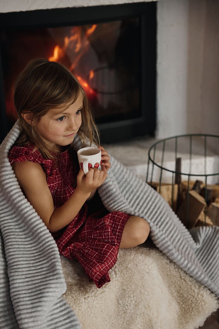 Une tasse de chocolat chaud ou de glögg fait partie intégrante de la fête de Noël suédoise. Ici, vous voyez une fillette assise sur un canapé devant la cheminée avec la tasse Children of the forest et la couverture Pleece rabattue, toutes deux de Design House Stockholm.