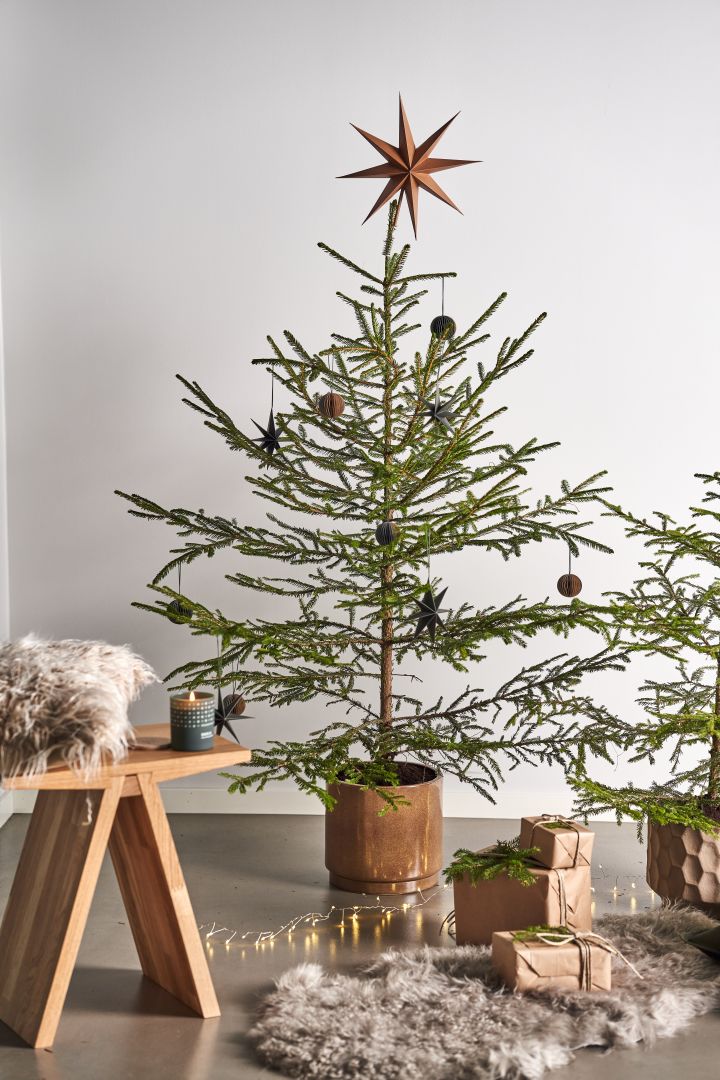 Décorez le sapin de Noël avec des décorations de sapin de Noël pour 2021 dans 4 styles différents selon les tendances Nest - Nurture, Share, Boost et Cultivate. Ici, vous voyez un tabouret Angle de Muubs avec une bougie parfumée Skandinavisk dans la senteur Forest à côté de paquets sur un tapis en peau de mouton. 