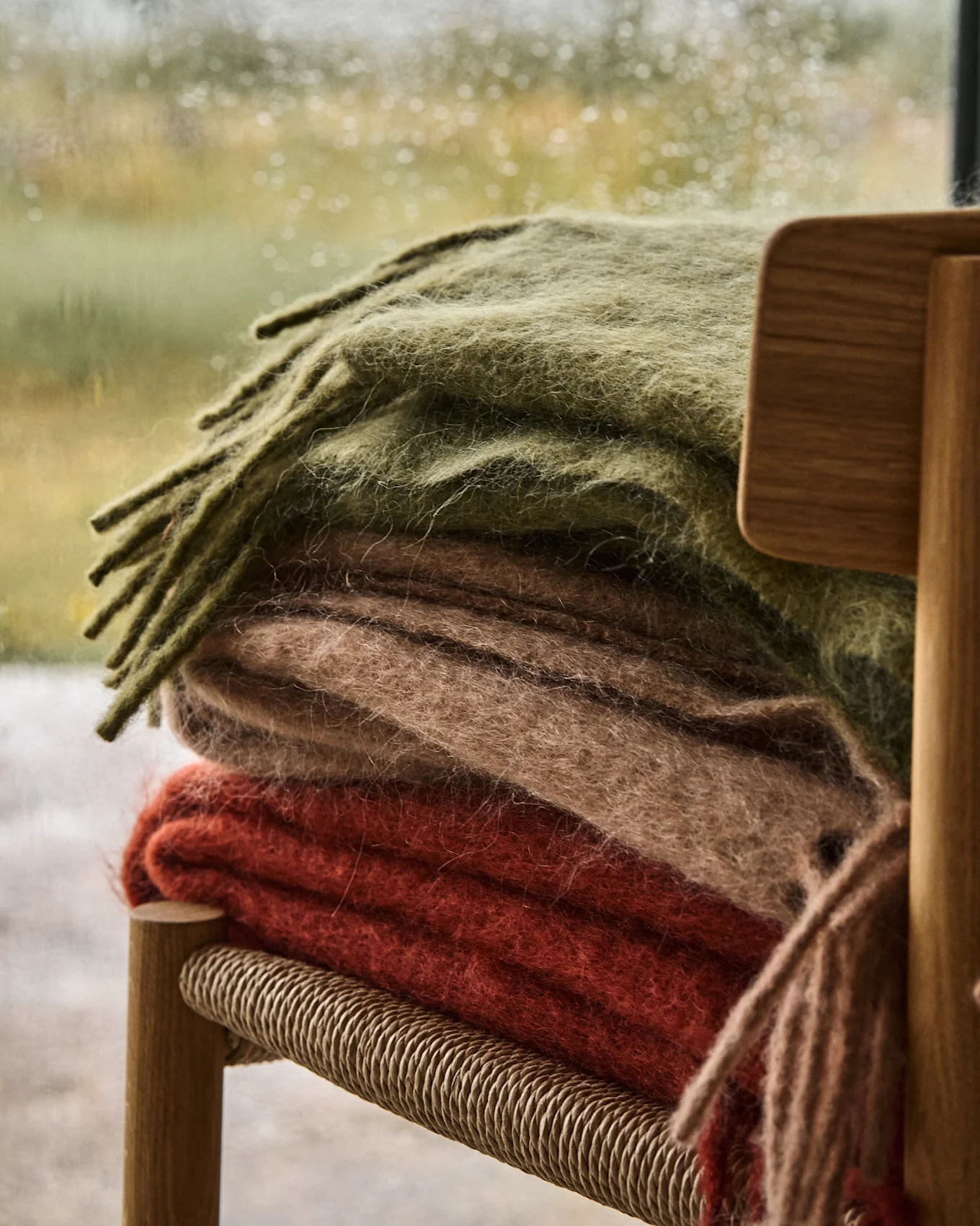 Pile de trois couvertures douces vert olive, beige et rouge-orange sur une chaise en bois, avec une fenêtre pluvieuse en arrière-plan.