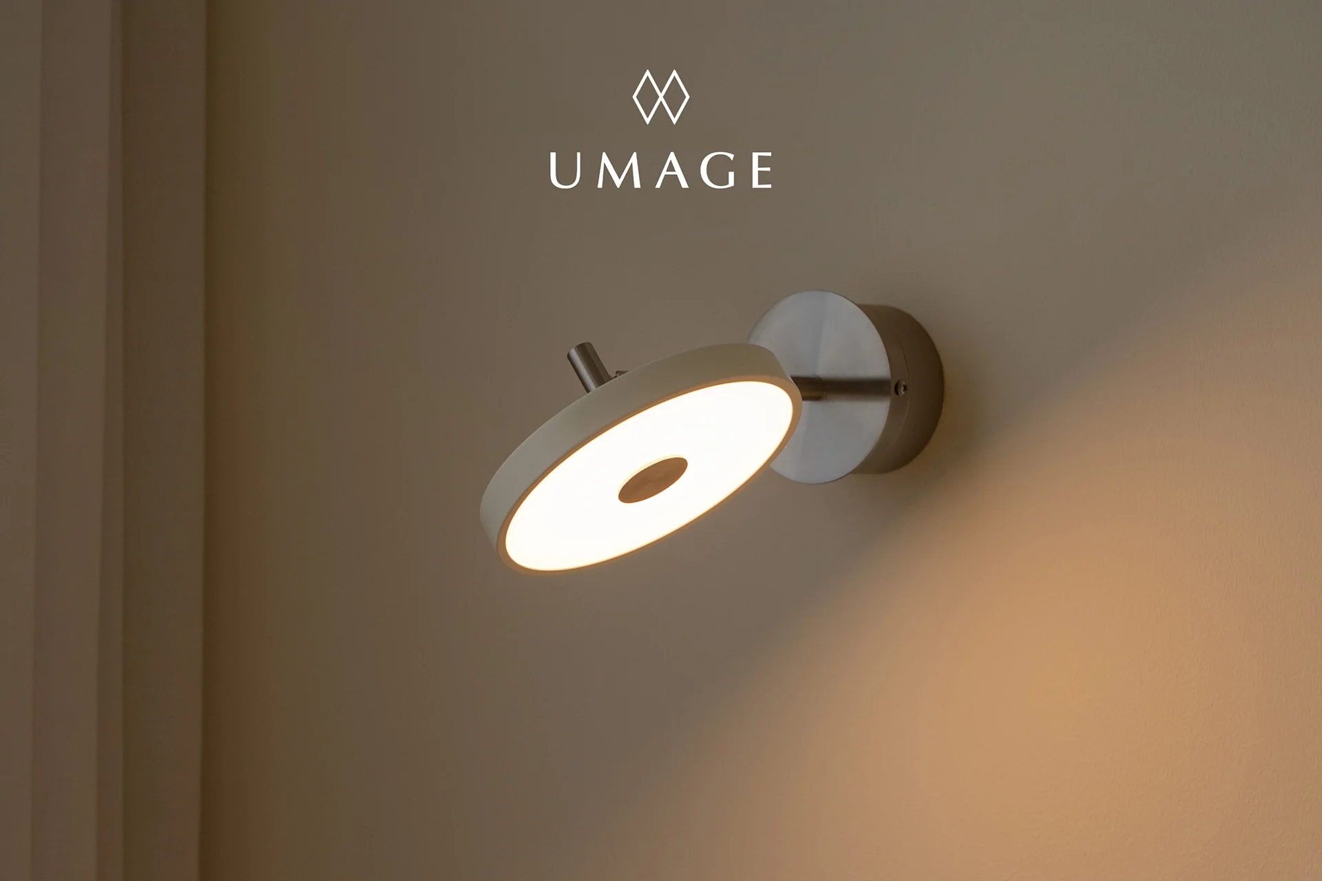 Une applique murale circulaire moderne avec une lumière chaude, montée sur un mur marron clair. Logo UMA au-dessus.