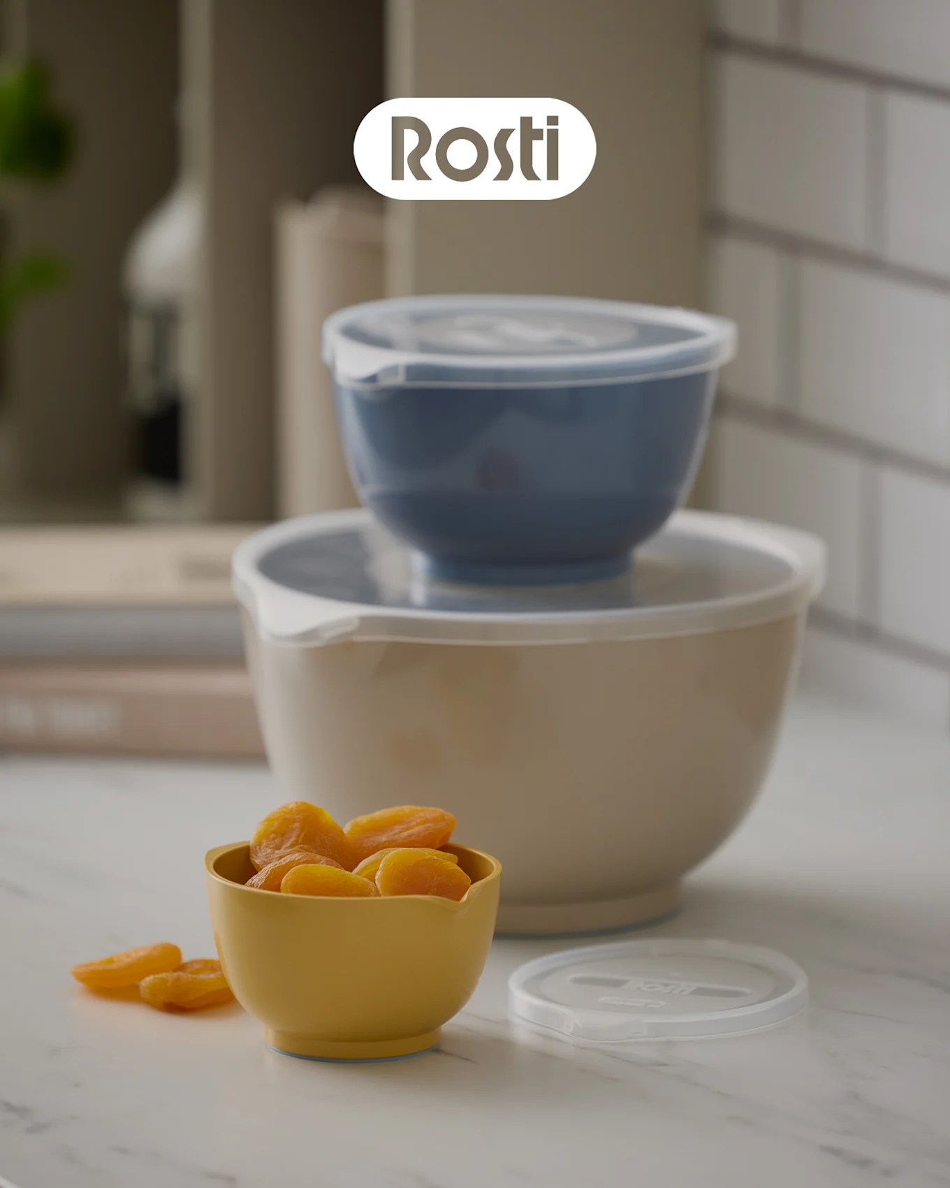 Trois bols Rosti sur un comptoir : un bol beige avec un bol bleu empilé dessus, les deux avec des couvercles, et un petit bol jaune rempli d'abricots secs.