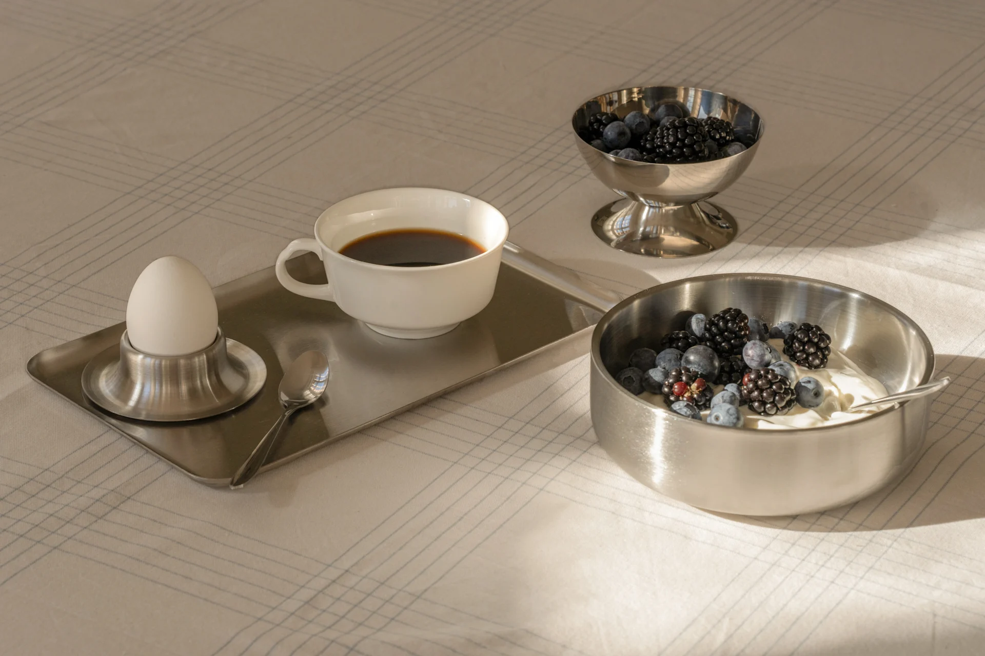 Petit-déjeuner avec café, œuf à la coque, yaourt et baies fraîches dans des bols argentés sur une nappe quadrillée.