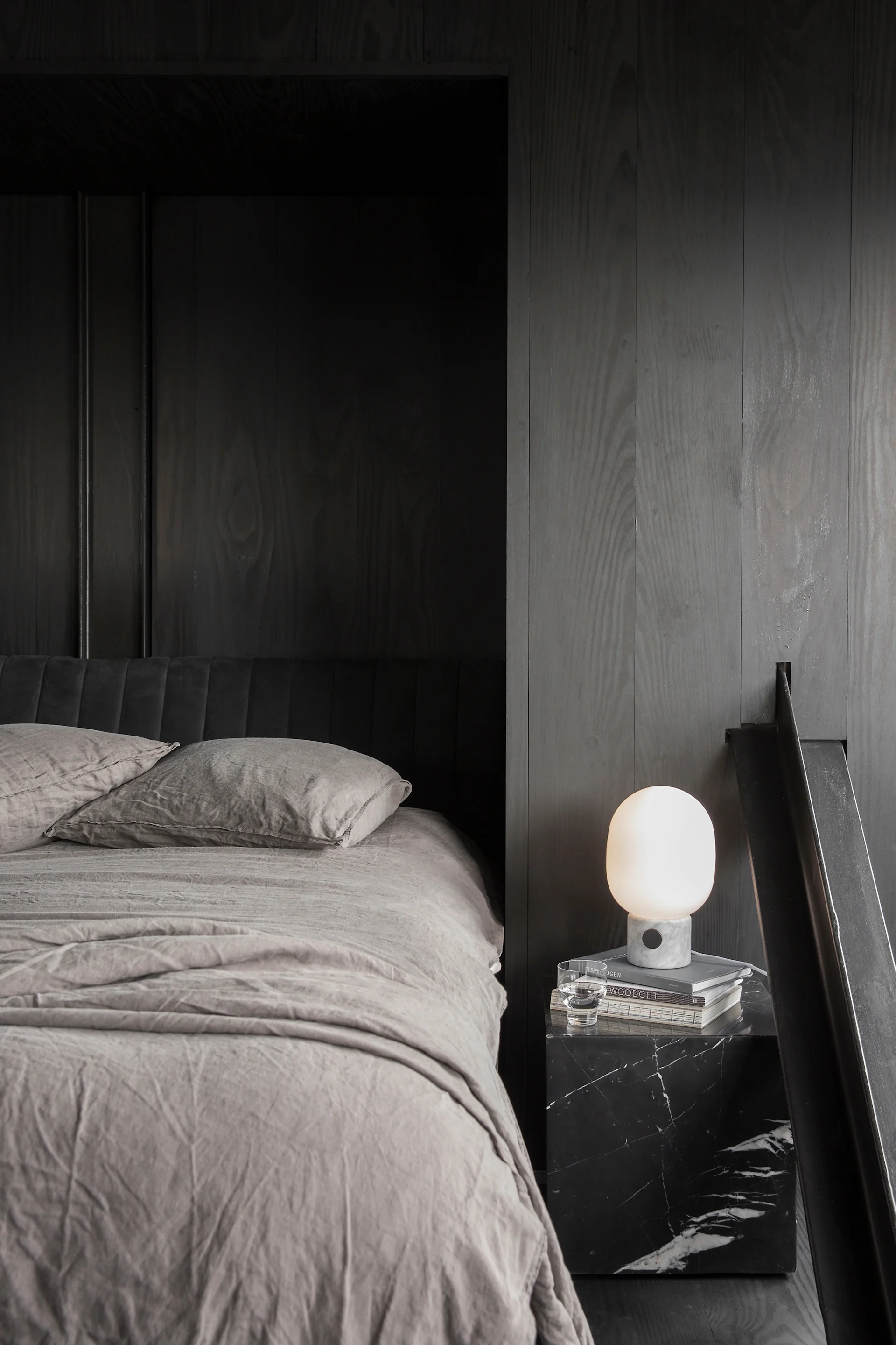 Une chambre moderne avec une literie en lin gris clair et une tête de lit foncée. Une table de chevet en marbre noir contient une lampe lumineuse, des livres et un verre d'eau.