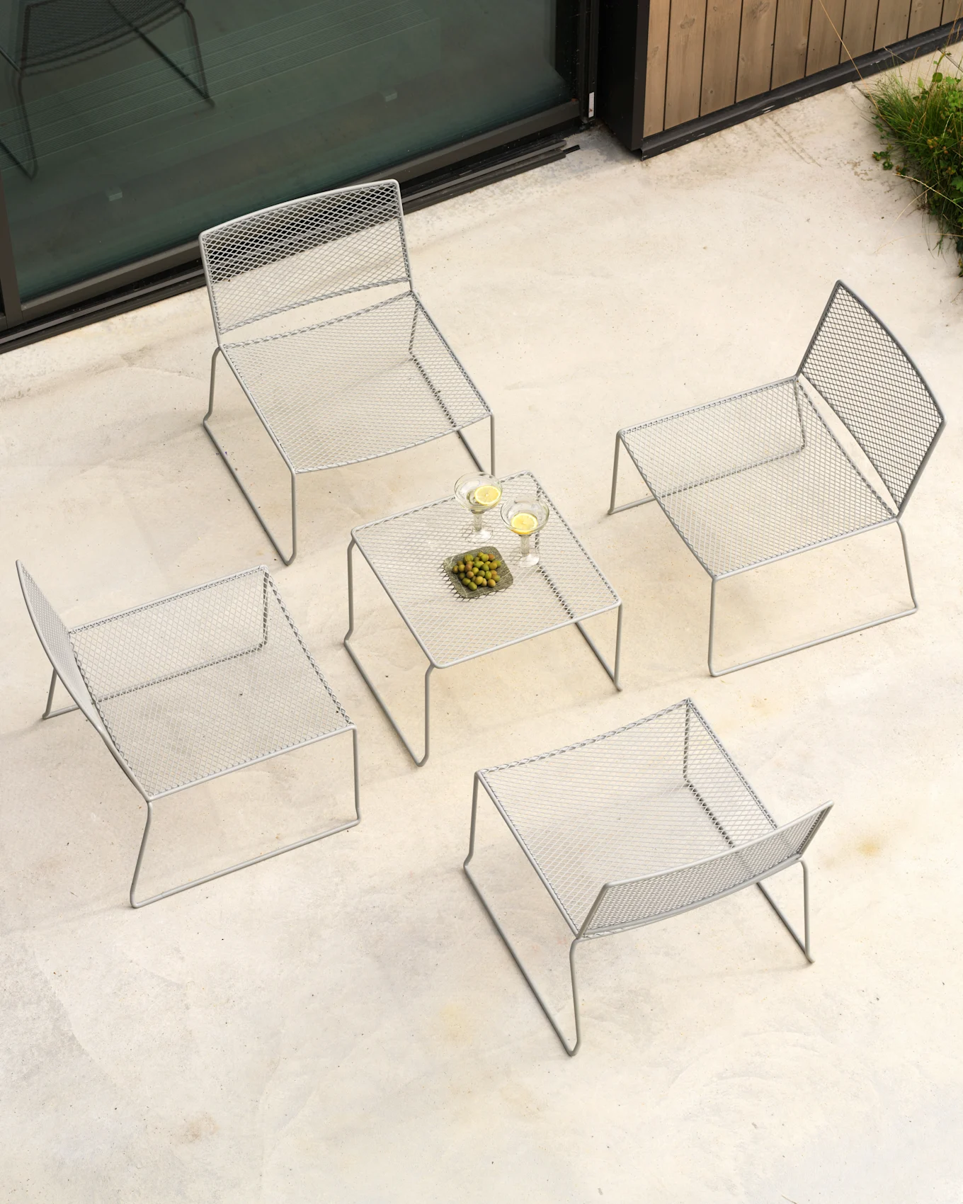 Vue aérienne d'un salon de patio extérieur avec quatre chaises en treillis métallique gris et une table basse assortie avec boissons et olives.