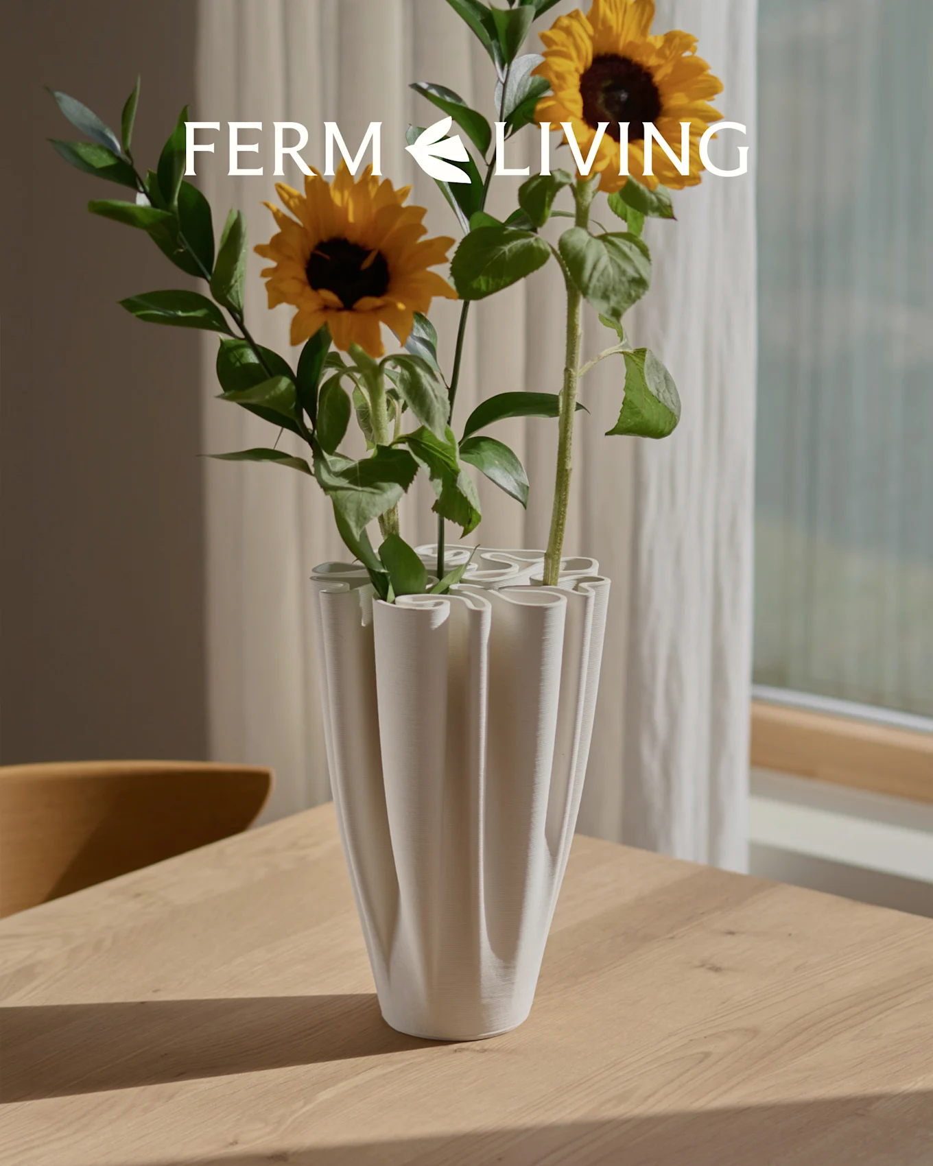 Tournesols dans un vase blanc, nervuré et ondulé sur une table en bois. Superposition du logo Ferm Living.