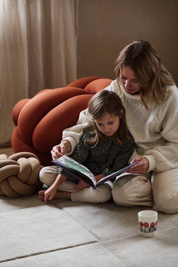 Pour les familles, la lecture d'une histoire de Noël suédoise fait partie intégrante de la fête de Noël. Ici, vous voyez une mère et sa fille assises sur le tapis Basket et feuilletant un livre, entourées d'un petit et d'un grand coussin Knot, tous de Design House Stockholm.