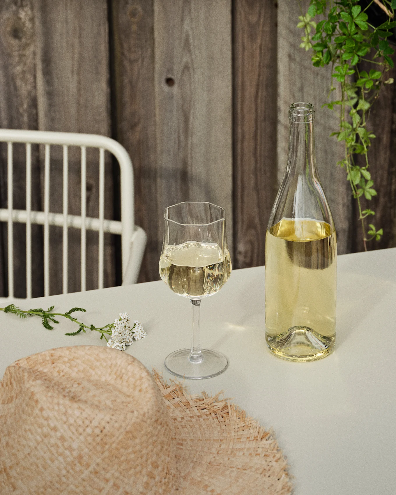 Une bouteille et un verre de vin blanc sur une table avec un chapeau de paille et des fleurs blanches, devant un mur en bois et des vignes vertes.
