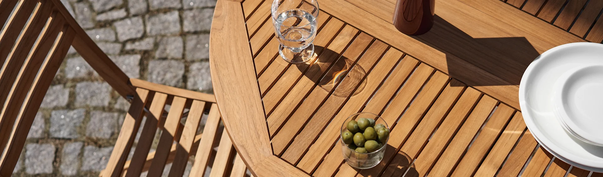 Vue plongeante d'une table de patio en lattes de bois avec un verre d'eau, un bol d'olives vertes et des assiettes blanches au soleil.