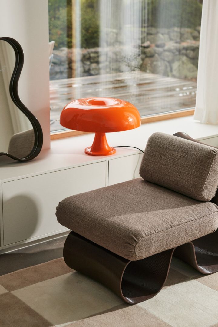 La lampe Nessino en orange avec son abat-jour en forme de beignet se trouve devant une fenêtre avec un miroir à bord incurvé et une chaise longue assortie. 