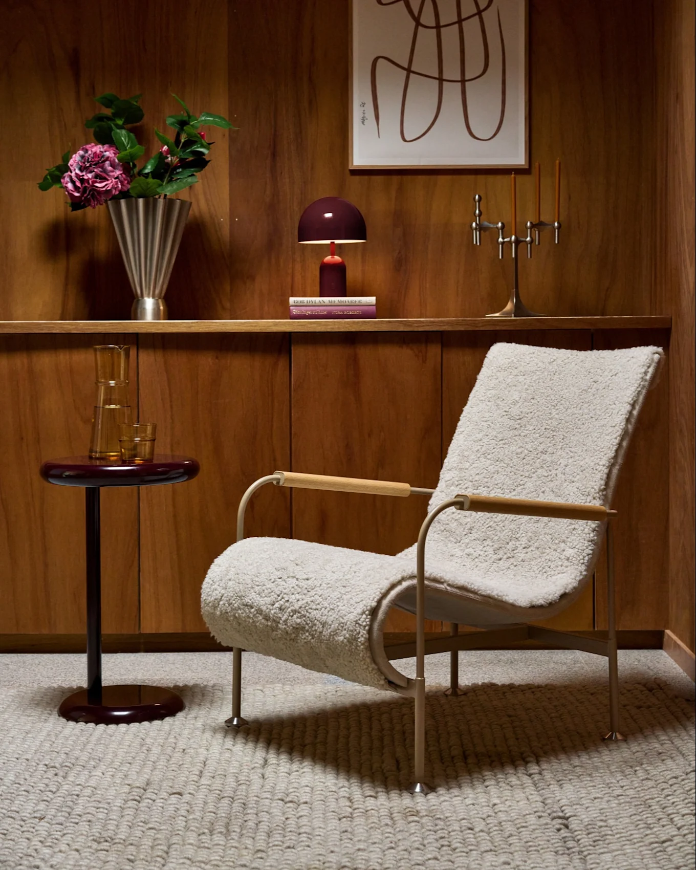 Pièce lambrissée avec fauteuil moelleux, table d'appoint rouge, étagère avec vase, lampe, candélabre et art abstrait.