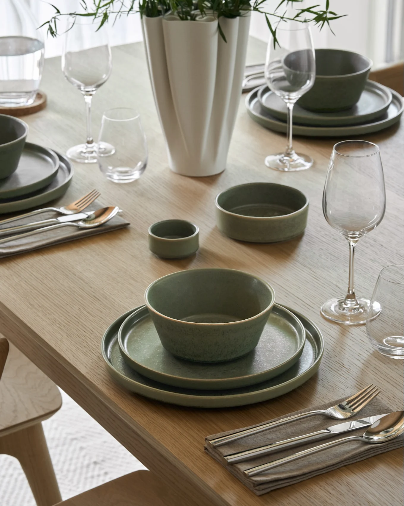 Table à manger dressée avec des bols de soupe crémeuse, des assiettes assorties, des verres à vin et à eau, des couverts et un vase avec du feuillage vert.