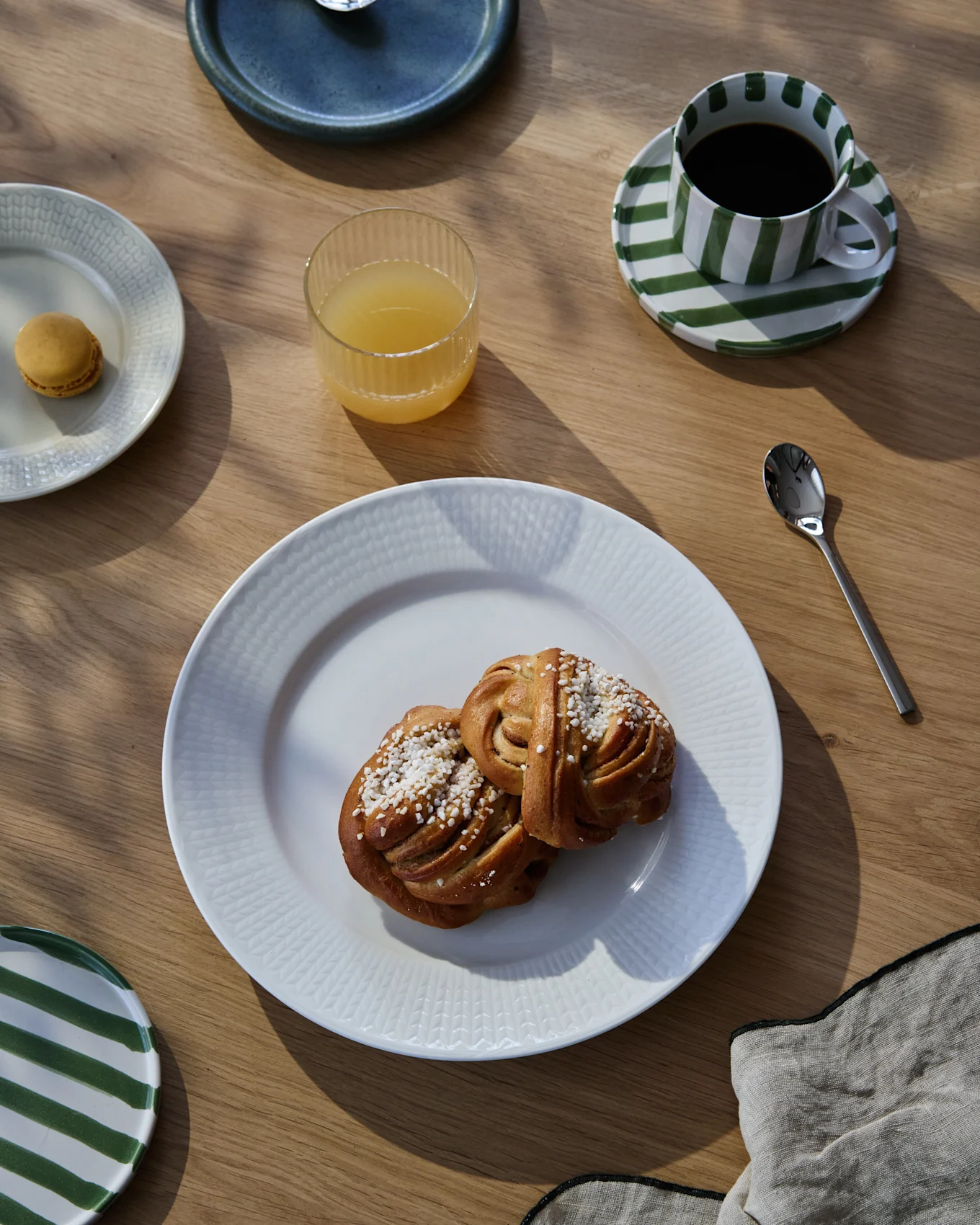 Un petit-déjeuner sur une table en bois avec deux roulés à la cannelle, du café dans une tasse rayée, du jus d'orange et un macaron.