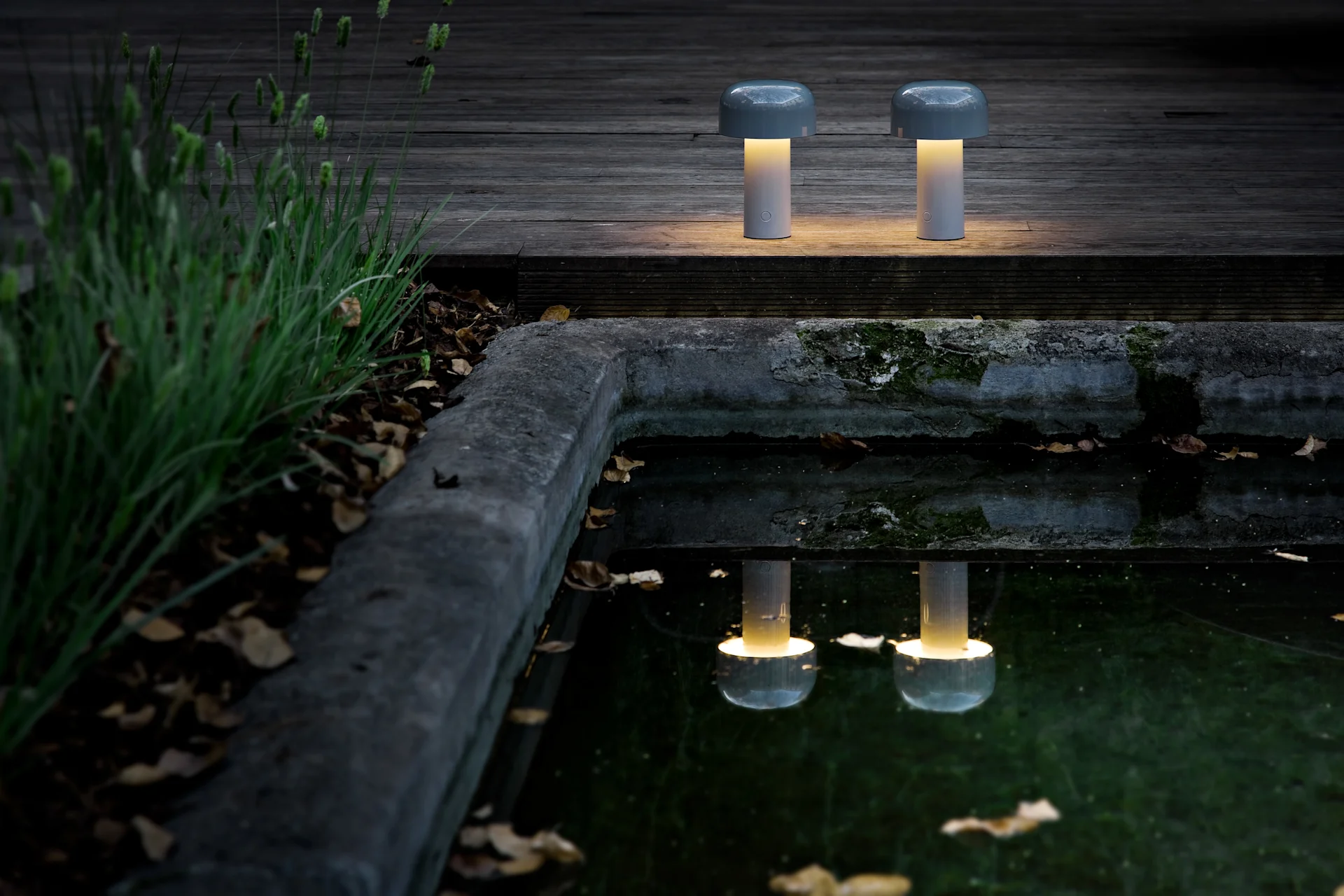 Deux lampes de table portables en forme de champignon avec une lumière jaune chaude sur une terrasse en bois sombre près d'un étang, reflétant leur éclat.