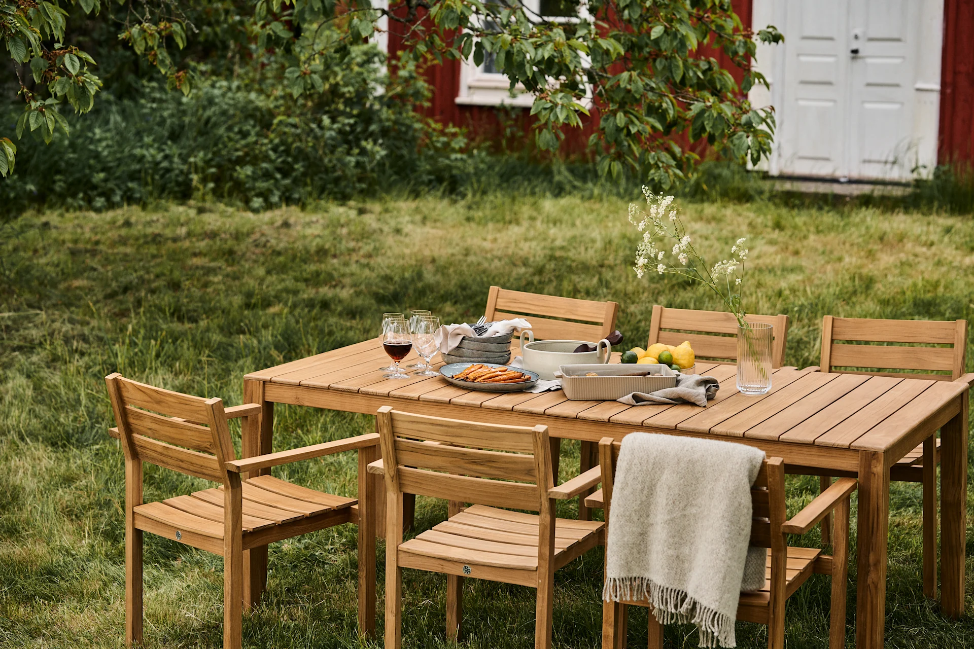 Ensemble de repas extérieur avec table en bois et six chaises sur une pelouse, garni de nourriture, de boissons et d'un plaid, avec une maison rouge en arrière-plan.