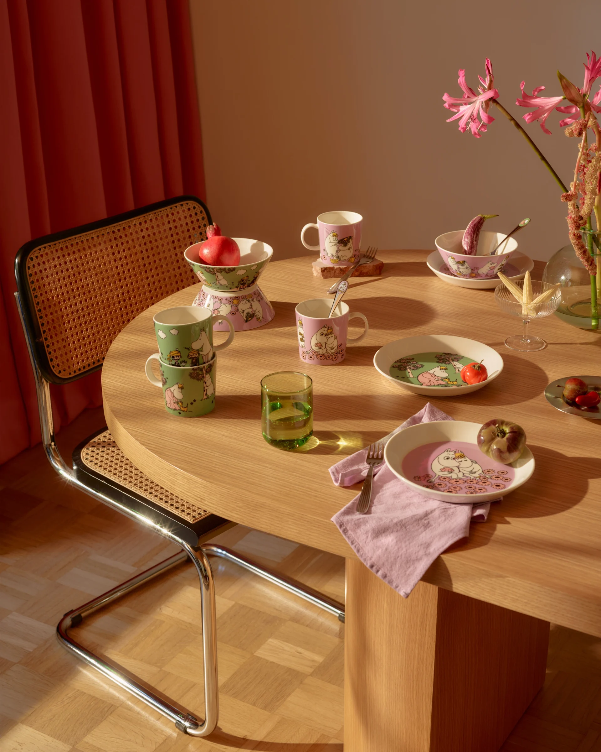 Table à manger avec vaisselle à thème Moomin rose et verte, fruits frais, un verre vert et des fleurs roses.