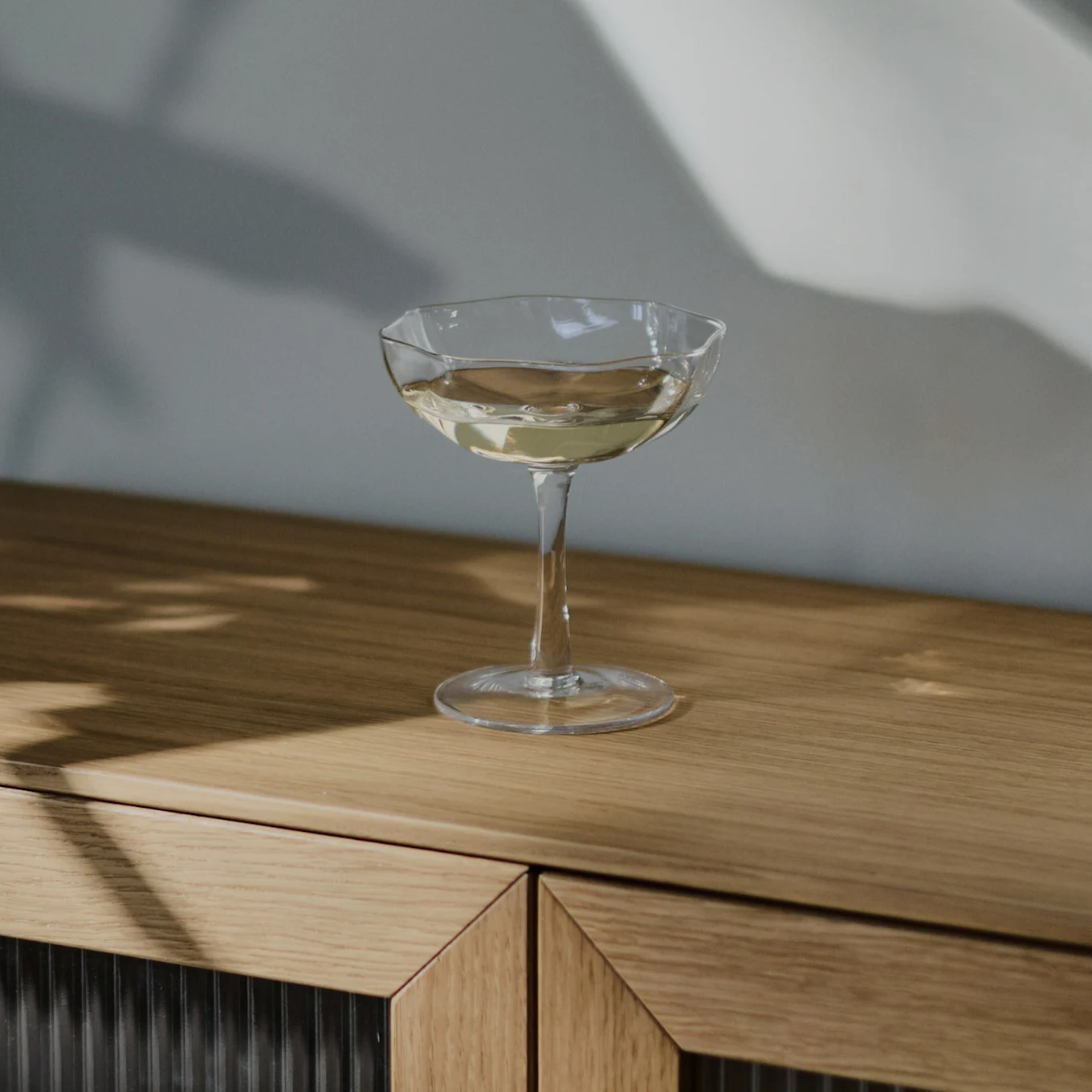 Un verre à coupe transparent rempli d'une boisson claire repose sur un meuble en bois clair, avec des ombres sur le mur.