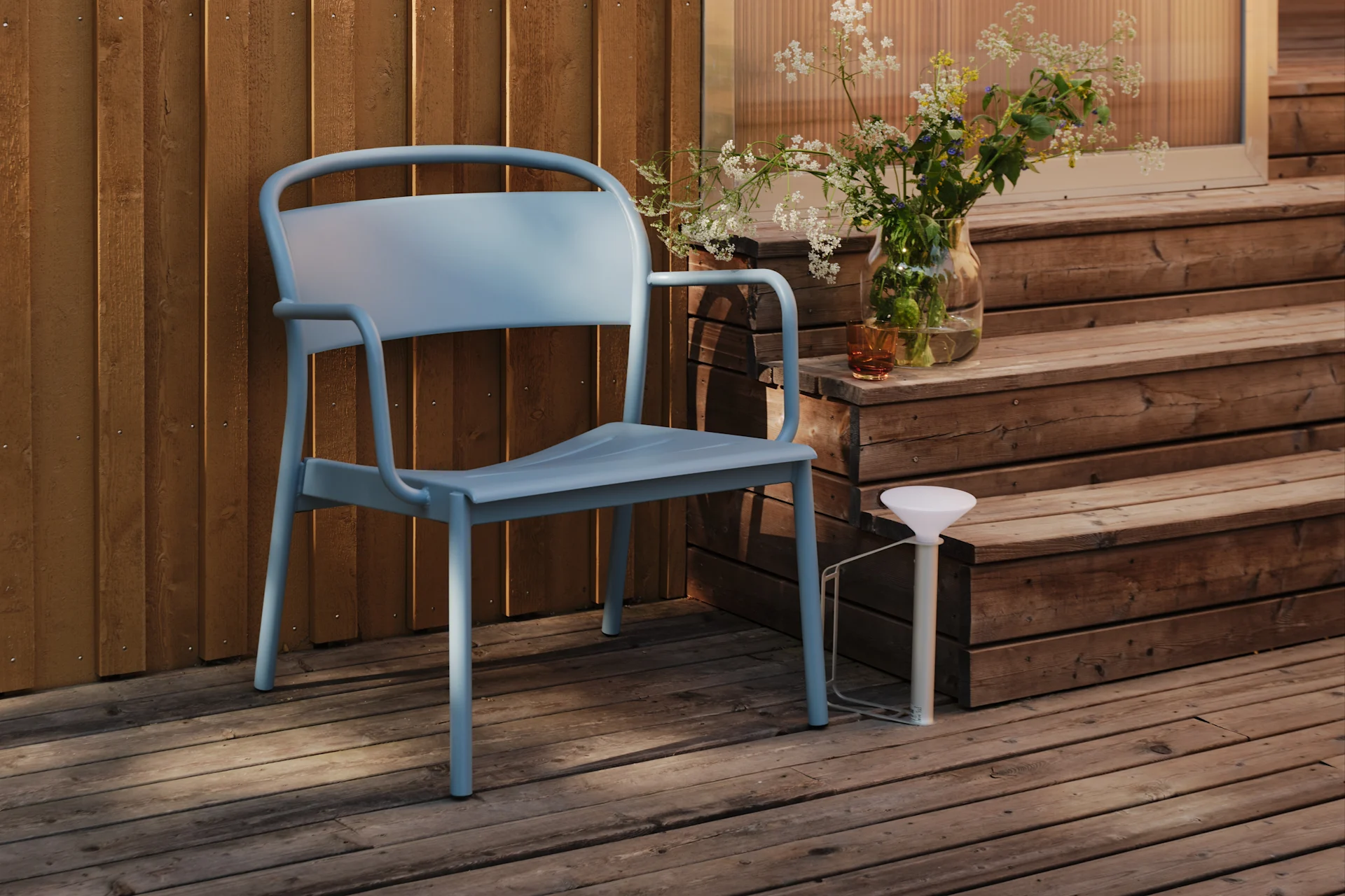 Une chaise en métal bleu clair est posée sur une terrasse en bois à côté d'un mur en bois, avec un vase de fleurs sauvages sur une marche.