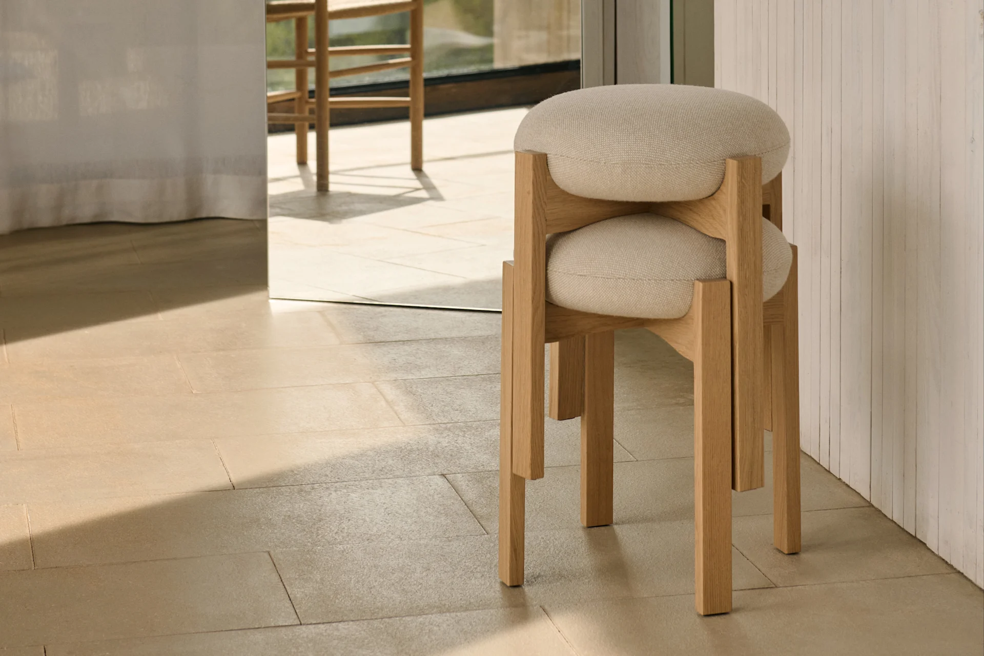 Deux tabourets minimalistes en bois clair avec des sièges ronds rembourrés empilés sur des carreaux clairs, avec une vue extérieure ensoleillée à travers une grande fenêtre.