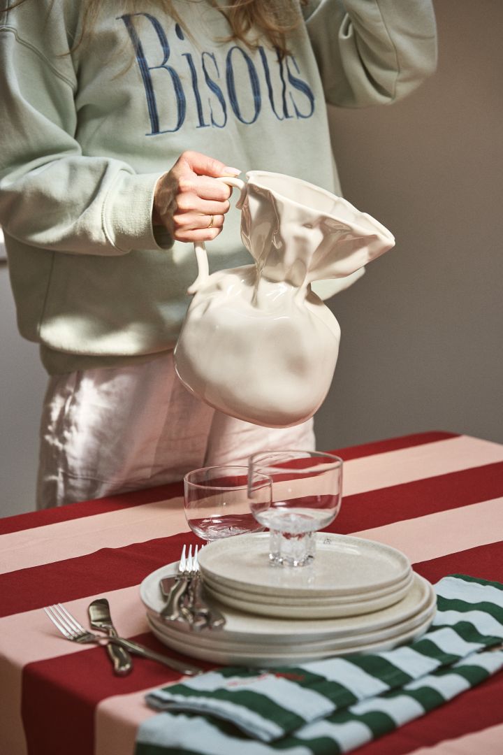 Personne tenant le vase Crumple en blanc de Byon à une table ornée d'une nappe rayée rose et bordeaux. Le vase Crumple a la forme d'un sac en papier froissé et est fabriqué en céramique émaillée.