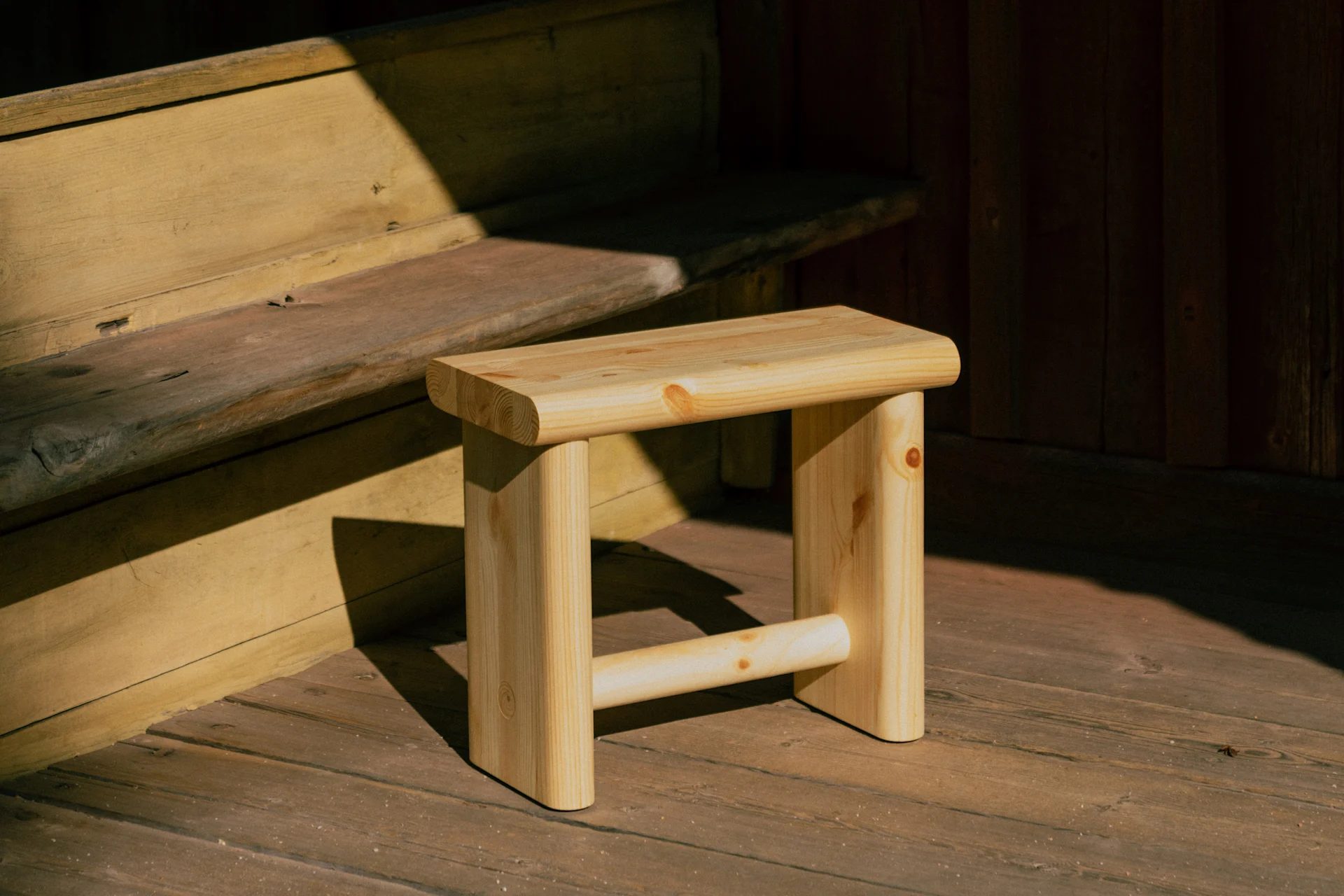 Un tabouret en bois clair est posé sur un plancher en bois, partiellement éclairé par le soleil, à côté de marches en bois et d'un mur en bois foncé.