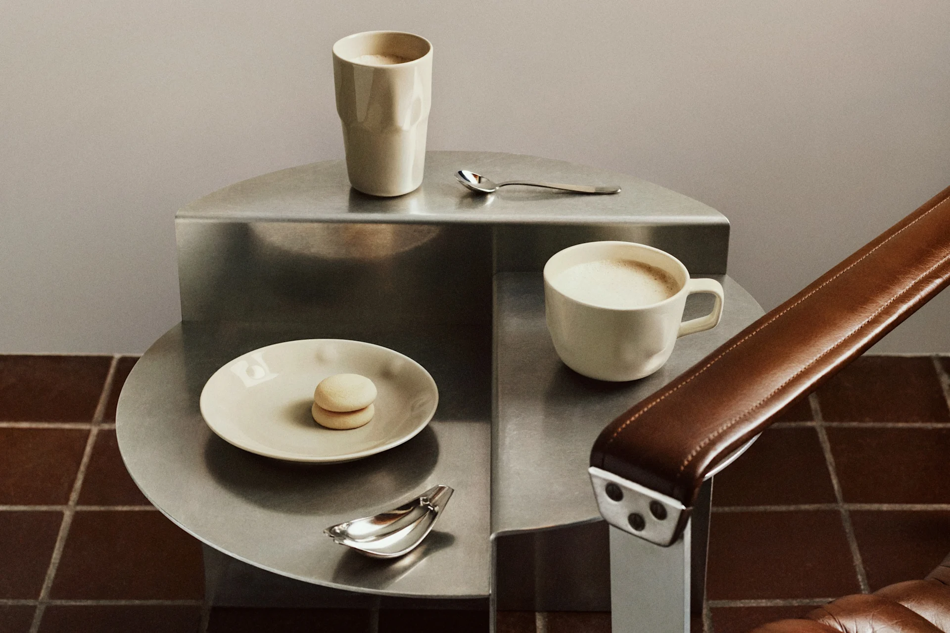 Deux tasses à café claires, un macaron et des cuillères sur une table d'appoint métallique étagée à côté d'un fauteuil en cuir marron.
