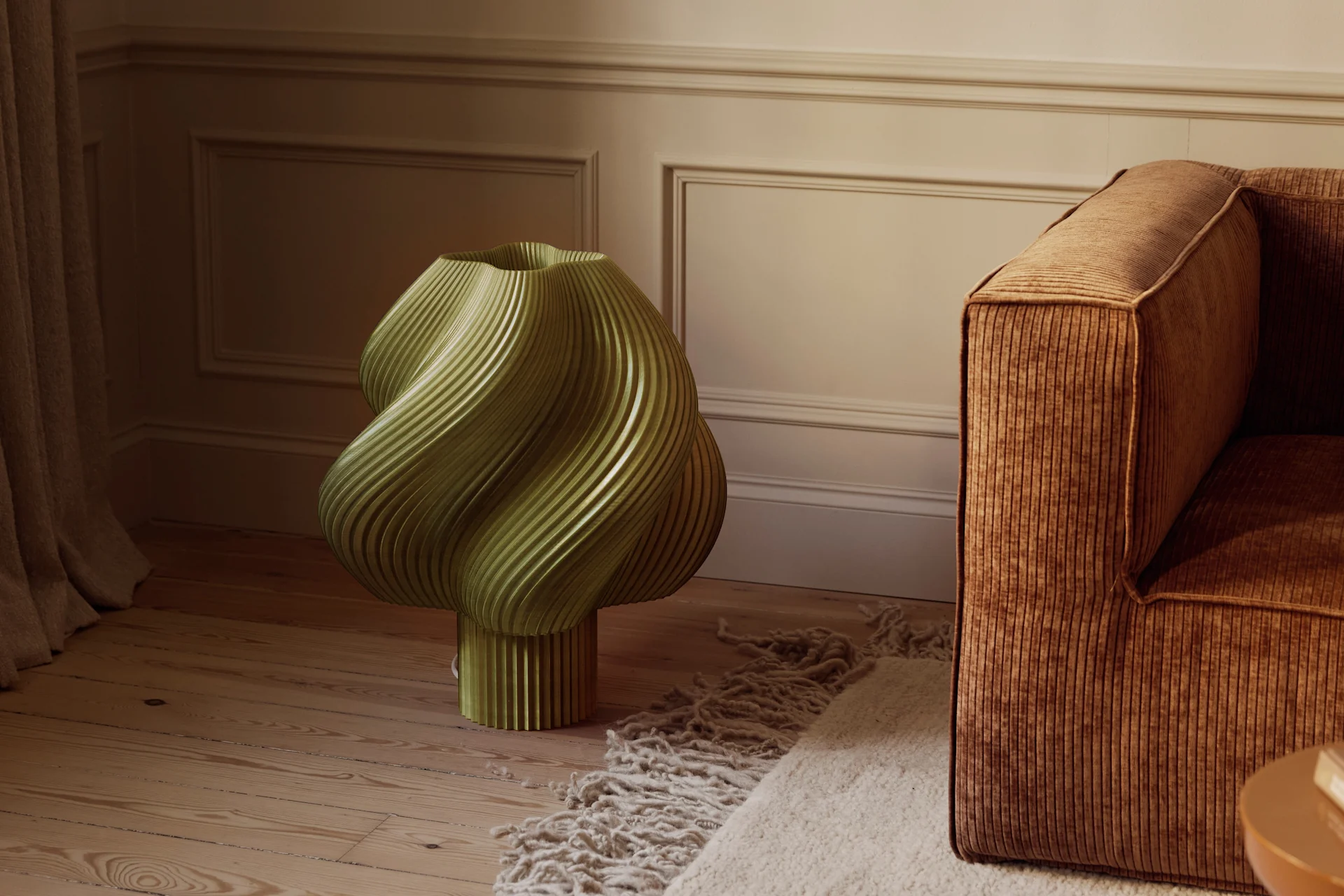 Un objet sculptural vert olive cannelé sur un parquet en bois, à côté d'un canapé en velours côtelé marron et d'un tapis texturé, dans une pièce aux murs moulurés.