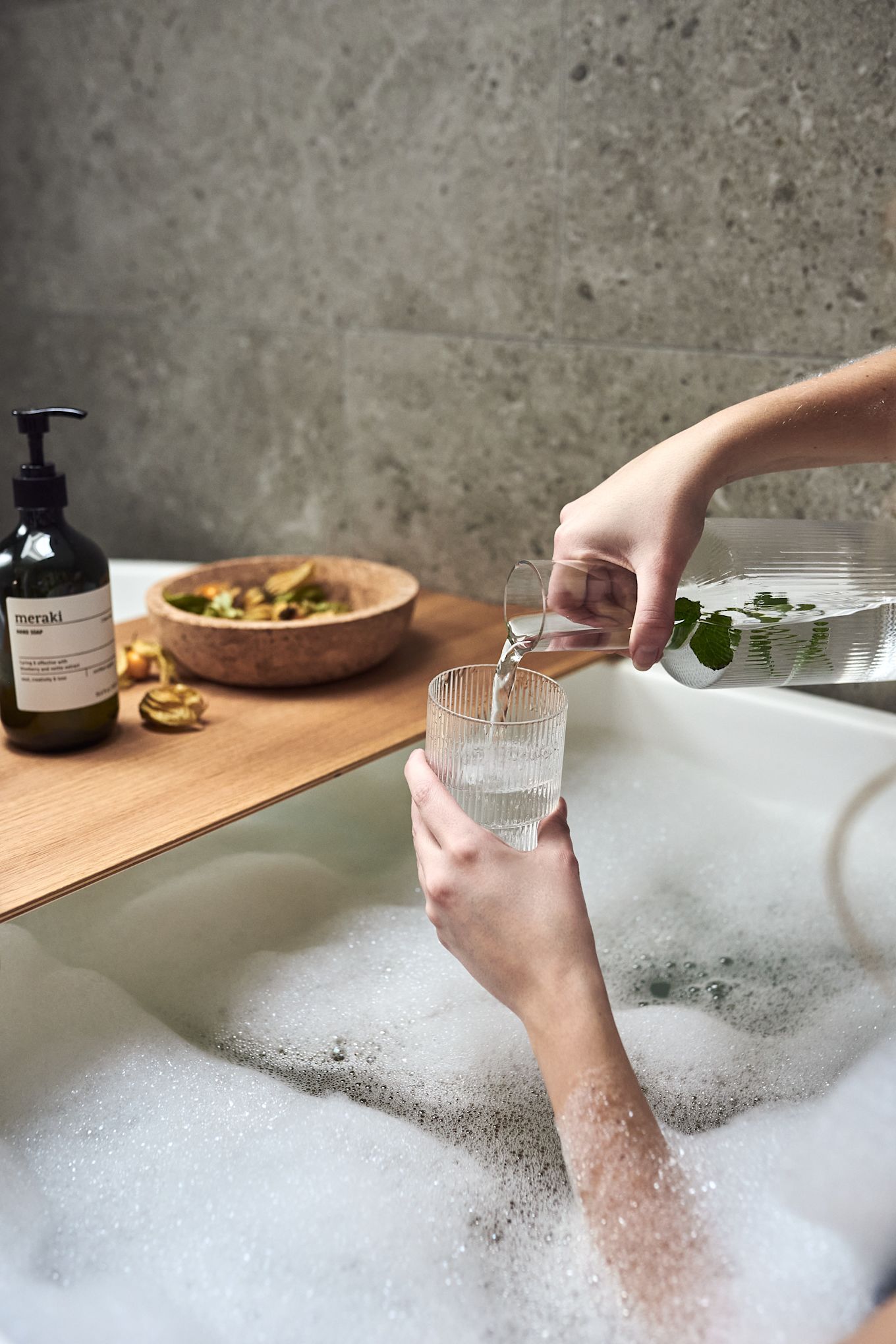 Des mains versent de l'eau dans un verre Ripple de Ferm Living dans le cadre d'une soirée spa à domicile avec un bain moussant chaud. 