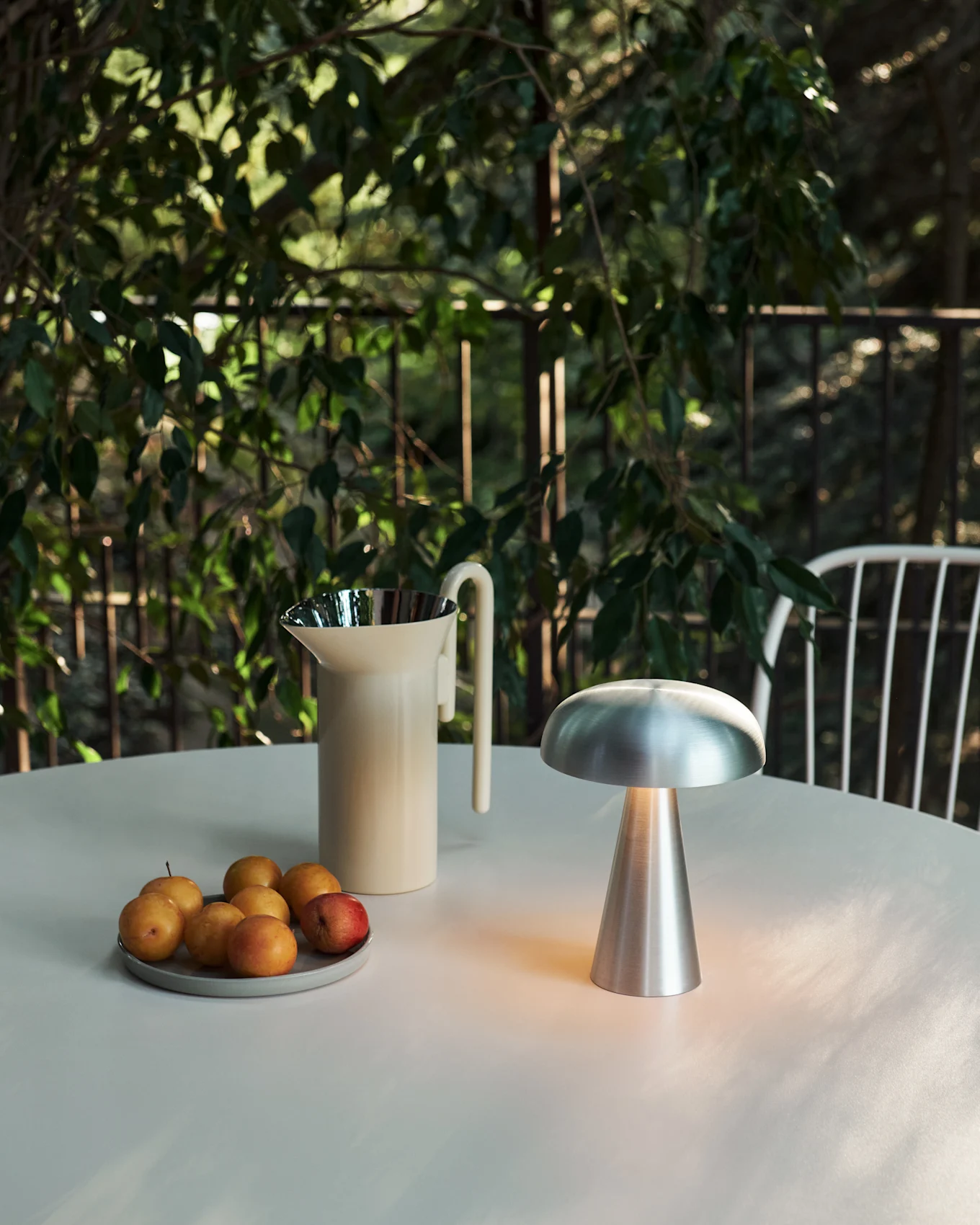 Table d'extérieur avec lampe champignon argentée lumineuse, carafe crème et assiette de prunes, entourée de feuillage vert.