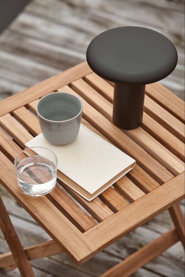 La lampe portative Takku d'Artemide est posée à l'extérieur sur une table en bois avec une tasse à café et un verre d'eau. 