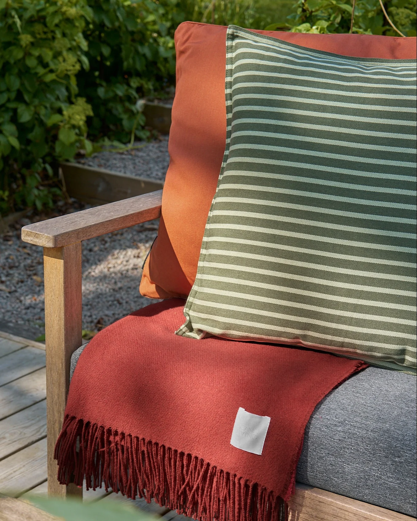 Chaise de jardin en bois avec coussin orange, oreiller rayé vert et blanc, et plaid frangé couleur terre cuite.