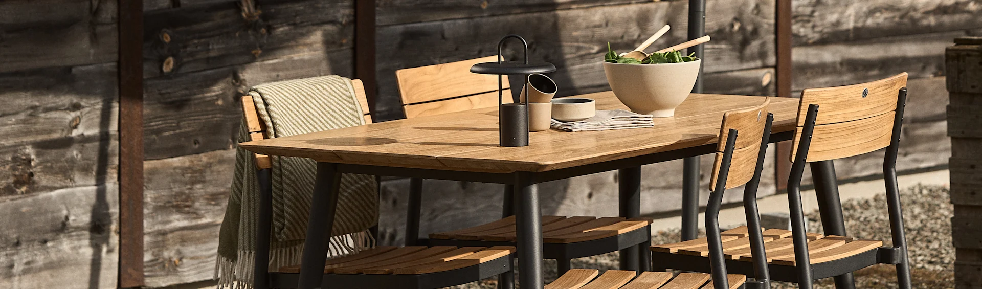 Ensemble de repas extérieur avec une table en bois, des chaises noires, une lanterne, un saladier et une couverture drapée.