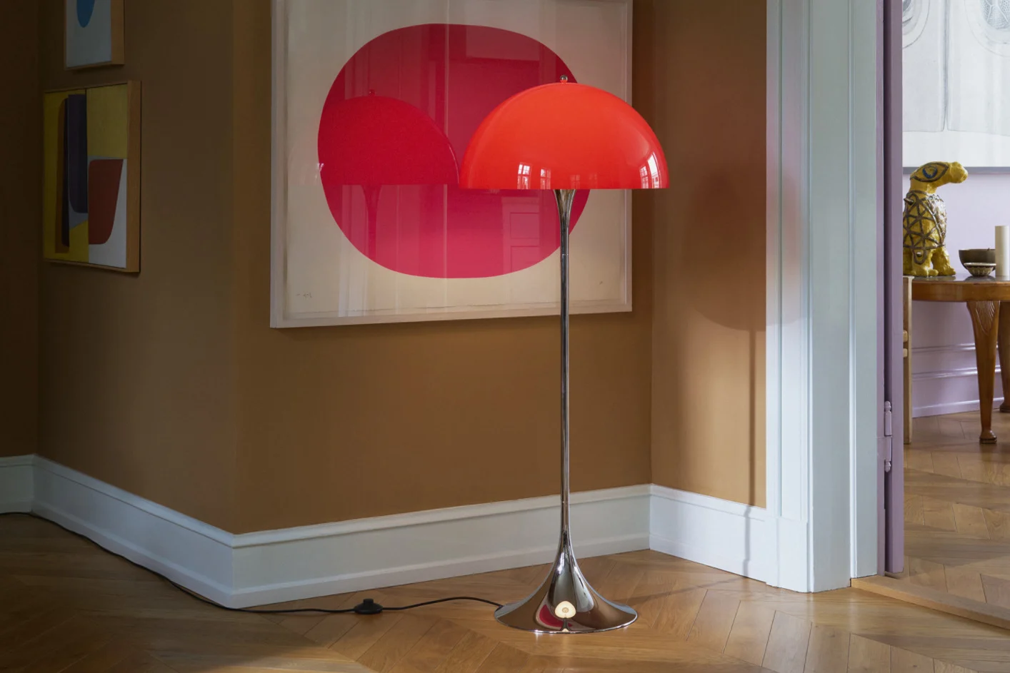 Un lampadaire rouge vif en forme de dôme avec un pied chromé sur un parquet dans une pièce aux murs marron et à l'art moderne.